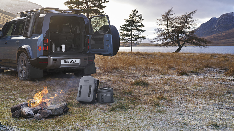 backpack and cool bag on the floor next to a camp fire, Defender vehicle in the background